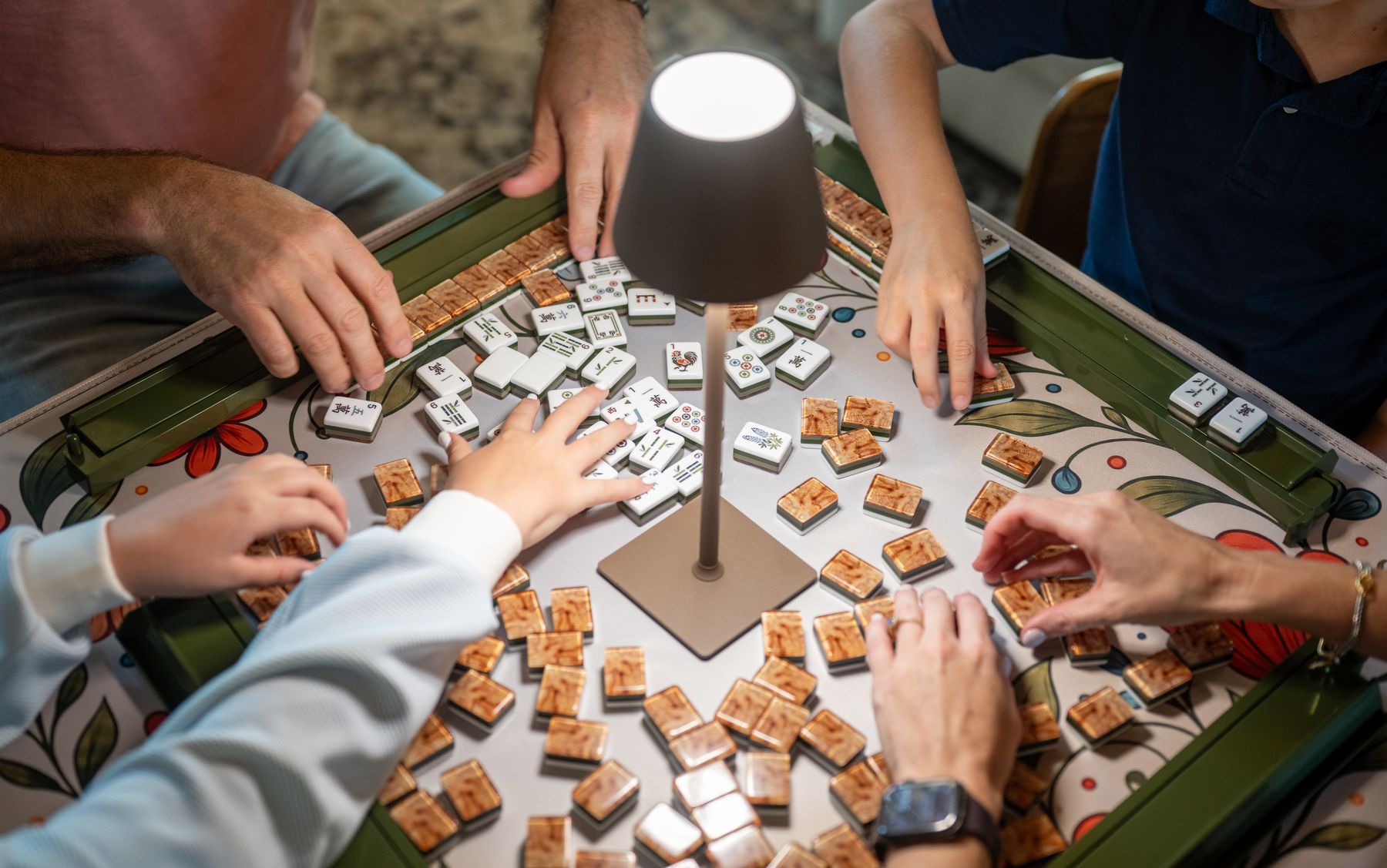 Hands shuffling tiles under a table lamp